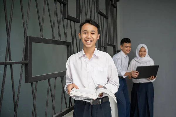En asiatisk tonåring i skoluniform ler mot kameran medan han bär på en bok — Stockfoto