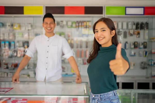 Krásná mladá žena s palci nahoru stojící na pozadí obchodu smartphone — Stock fotografie