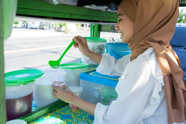 Uma menina bonita em um véu vende es campur usando uma colher para obter leite de coco — Fotografia de Stock