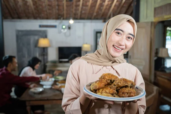 Tersenyum perempuan terselubung membawa sepiring ayam goreng dengan anggota keluarga makan bersama-sama — Stok Foto