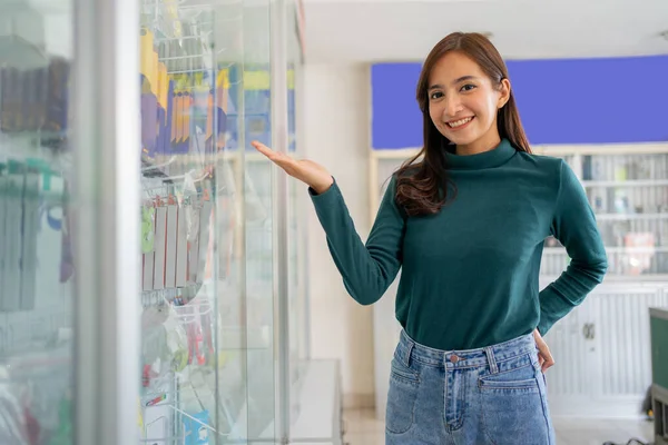 Piękna azjatycka kobieta uśmiecha się z gestem ręki oferuje coś w pobliżu telefon komórkowy akcesoria gablocie — Zdjęcie stockowe