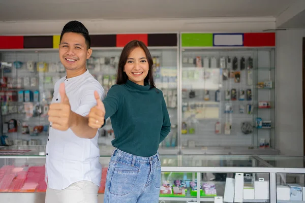 En stilig ung man och vacker ung kvinna med tummen upp stående på en smartphone butik — Stockfoto