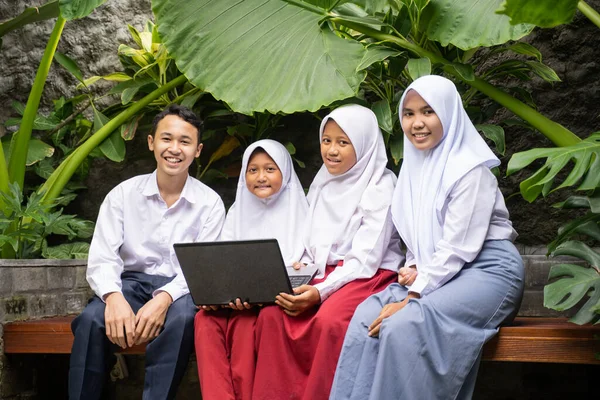Empat remaja berseragam sekolah tersenyum sambil duduk menggunakan laptop — Stok Foto