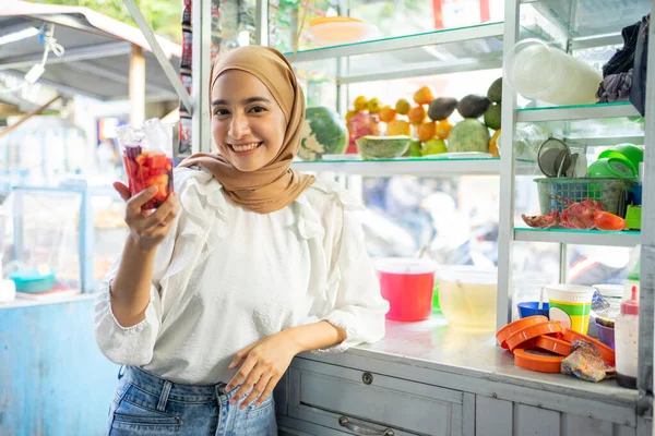 Una chica con una bufanda en la cabeza vendiendo hielo de frutas sonríe sosteniendo una taza de hielo de frutas — Foto de Stock