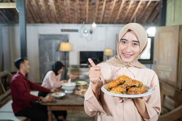 Wanita berkerudung membawa sepiring ayam goreng dengan jempol di latar belakang anggota keluarga makan bersama — Stok Foto