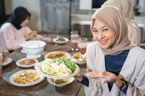 Una mujer velada sonríe a la cámara con gestos de mano mientras sostiene un plato de verduras — Foto de Stock