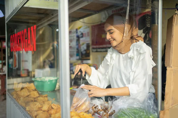 Une belle fille portant un hijab tout en utilisant des pinces choisit la nourriture frite que l'acheteur commande — Photo