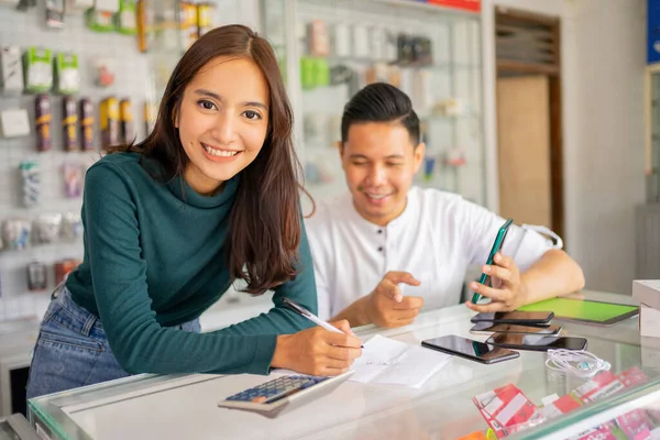 Hermosa mujer ayudante de tienda sonriendo mientras trabaja con un hombre que revisa las mercancías papel de nota con notas en el móvil —  Fotos de Stock