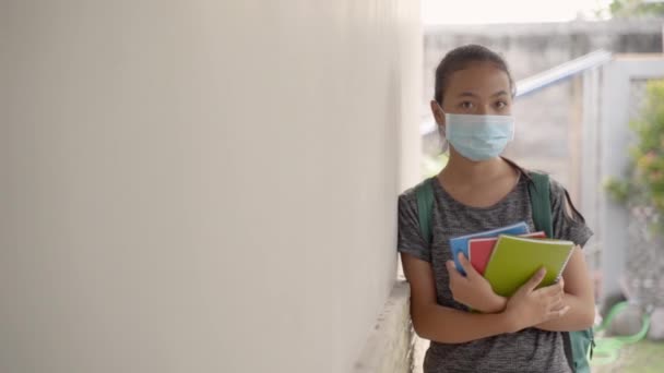 A teenage girl standing wearing a mask and backpack carrying a book — Stockvideo