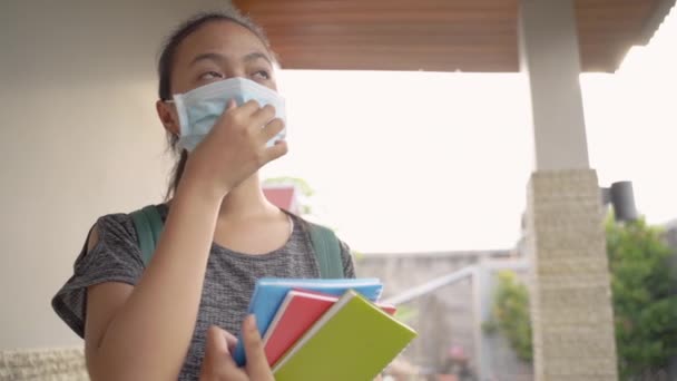 A teenage girl standing wearing a mask and backpack carrying a book — Stockvideo