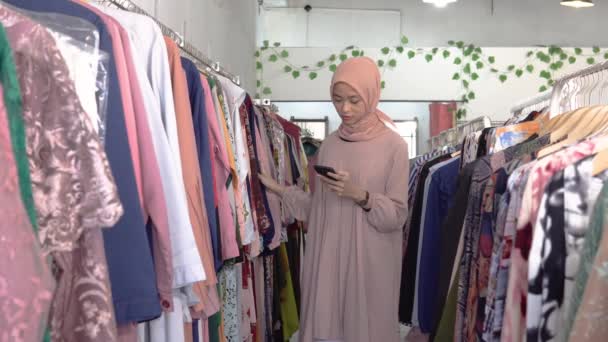 Ein schönes Mädchen im Hijab, das Kleidung wählt, während es ein Smartphone hält — Stockvideo