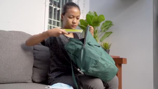 Genç bir Asyalı öğrenci okula gitmek için kitap ve sırt çantası hazırlarken maske takıyor. — Stok video