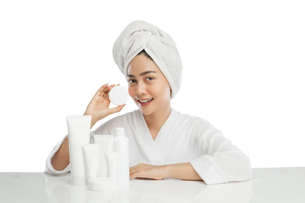 Smiling beautiful girl model with towel on head holding cream pot bottle with various skin care products on table — Fotografia de Stock