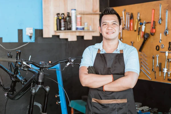 Close up of handsome bicycle mechanic in smiling apron holding bicycle with crossed hands — Stock fotografie