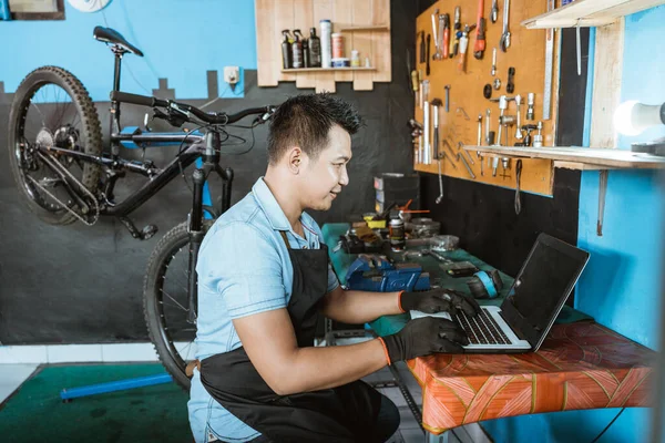 A bicycle mechanic in an apron wearing gloves sitting while using a laptop to look for spare parts —  Fotos de Stock
