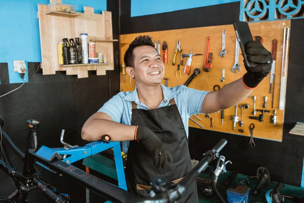 A smiling repairman in an apron selfie using a cellphone while repairing a bicycle — Fotografia de Stock