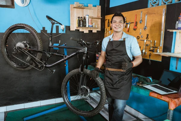 Bicycle mechanic in smiling apron holding bicycle with thumbs up while standing beside bicycle — Stock fotografie