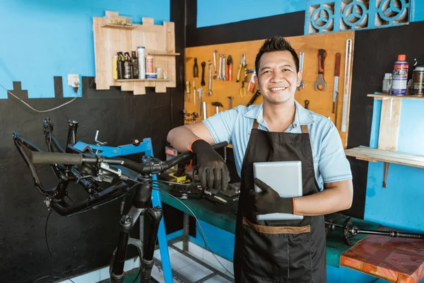 Portrait of a repairman in an apron leaning on a bicycle frame while holding a tablet — Φωτογραφία Αρχείου