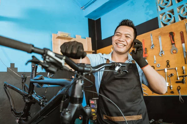 Retrato de un reparador en un delantal recibiendo una llamada por teléfono celular mientras reparaba una bicicleta — Foto de Stock