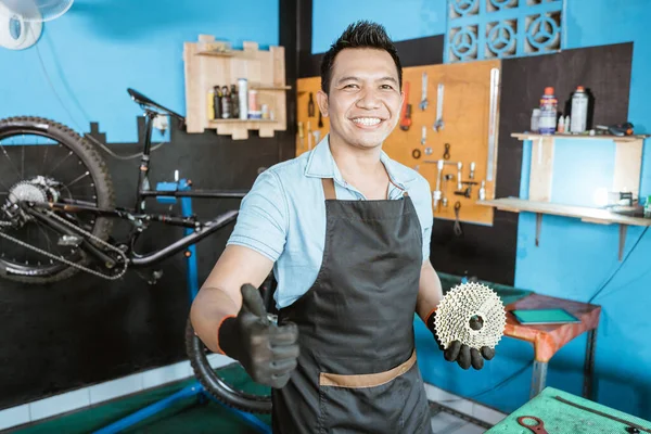 Handsome bicycle mechanic in smiling apron holding freewheel with thumb — Stock fotografie