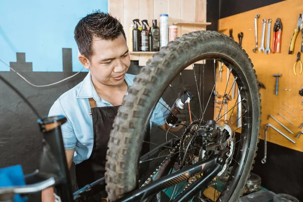 Mecánico de bicicleta guapo en delantal usa guantes mientras sostiene la botella mientras usa spray lubricante —  Fotos de Stock