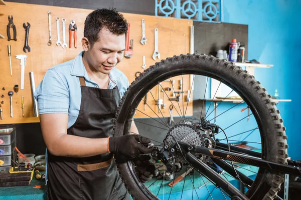 Un mecánico de bicicleta en el delantal sostiene el mecanismo trasero mientras soluciona problemas —  Fotos de Stock