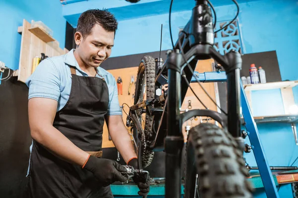 Mecánico de bicicleta guapo en un delantal con guantes al instalar pedales de bicicleta —  Fotos de Stock