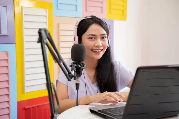 Cheerful girl looking at camera while using laptop and wearing headphones while talking into microphone during podcast — Stock fotografie