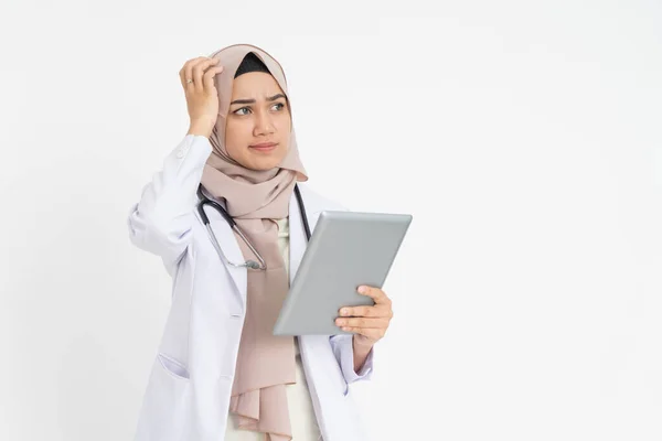 Velato medico donna che indossa uniforme tuta bianca confuso mentre guardando pad schermo con mano graffiante testa — Foto Stock