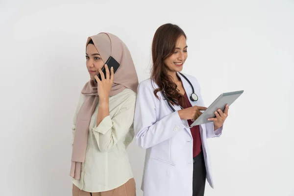 Allvarlig beslöjad kvinna ringer ett samtal med mobiltelefon och vacker kvinnlig läkare med hjälp av tablett med rygg till rygg pose — Stockfoto