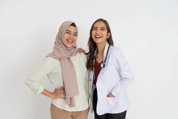 Kobieta w chustce i piękna kobieta lekarz śmieją się razem patrząc w aparat obejmujący pozę — Zdjęcie stockowe