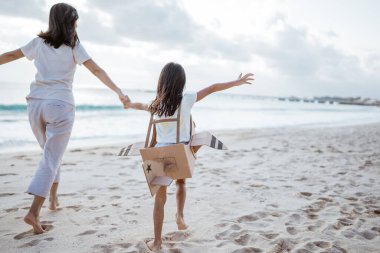 Anne kız el ele tutuşup karton uçakla koşuyor.