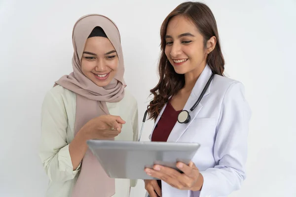 Usmívající se zahalená žena s ukazující rukou a šťastná krásná žena lékař při použití tablet spolu — Stock fotografie