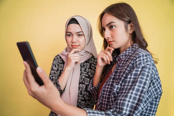 Eine Frau mit Kopftuch und eine Frau mit langen Haaren, die beim gemeinsamen Benutzen eines Smartphones ernsthaft auf den Bildschirm eines Mobiltelefons schaut — Stockfoto