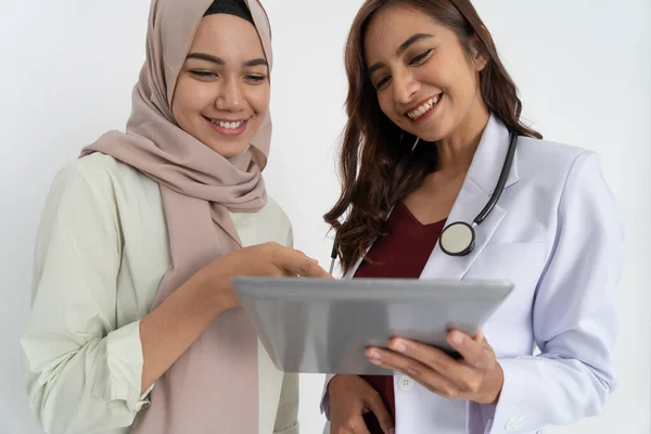 Sonriente mujer asiática en pañuelo de cabeza con las manos apuntando a la pantalla de la tableta y feliz hermosa doctora mientras usa la tableta juntos —  Fotos de Stock