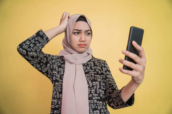 young woman in hijab confused while looking at smartphone screen with hand scratching head