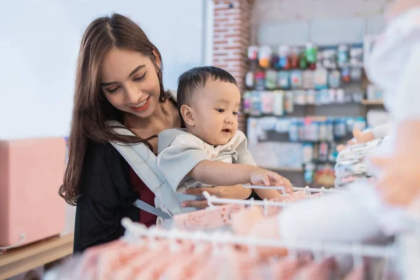 Madre recogiendo algún producto en la tienda de bebés mientras lleva a su hijo — Foto de Stock