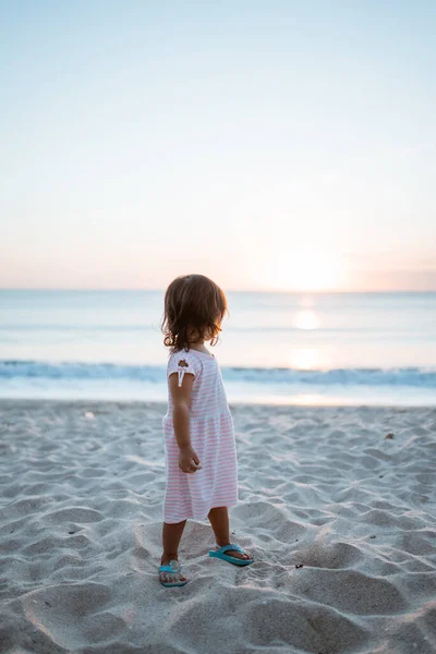 Κορίτσι από πίσω και ένα λευκό φόρεμα στέκεται σε μια καλοκαιρινή παραλία κοιτάζοντας το ηλιοβασίλεμα — Φωτογραφία Αρχείου
