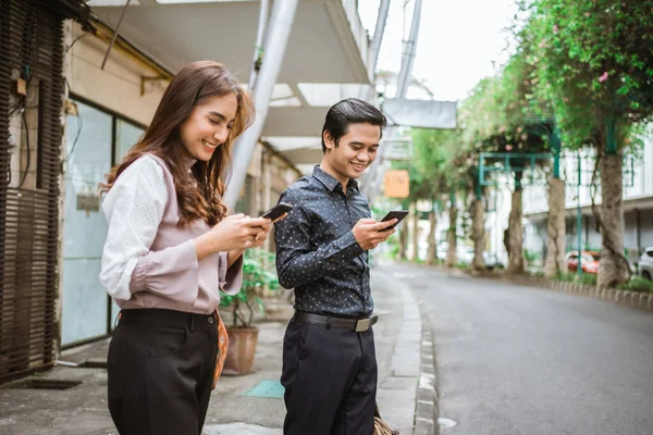 Mann und Frau stehen lächelnd auf dem Bürgersteig und benutzen ihr Handy — Stockfoto
