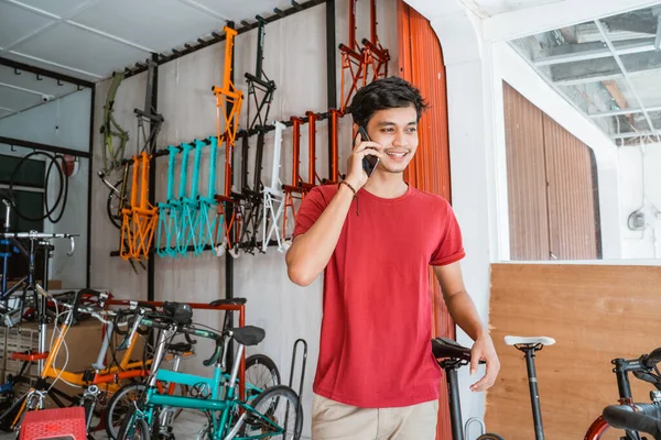 Hombre usando un teléfono móvil para hacer una llamada mientras sostiene una bicicleta nueva en la tienda de bicicletas —  Fotos de Stock
