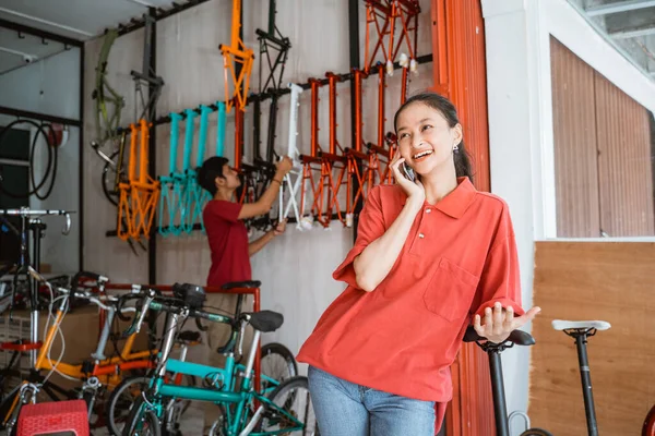 Mujer sonriendo felizmente mientras chatea usando un teléfono móvil en una tienda de bicicletas — Foto de Stock