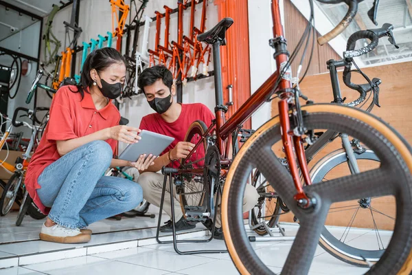 Majitel firmy a mechanik v masce zkontrolujte nové díly jízdních kol pro zákazníka pomocí tabletu — Stock fotografie