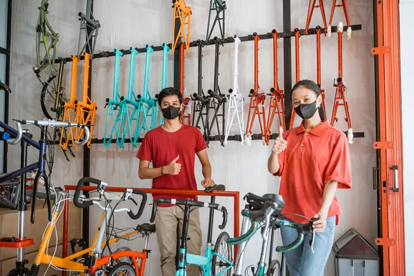 Hombre y mujer trabajador en máscara negra con los pulgares hacia arriba al sostener y preparar bicicletas — Foto de Stock