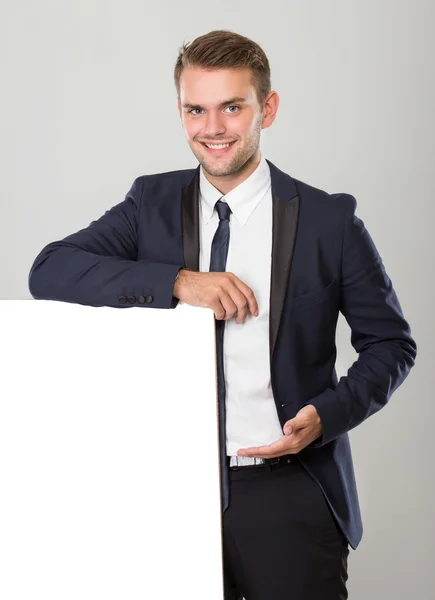 Joven hombre de negocios en traje negro apoyado en el signo blanco en blanco — Foto de Stock