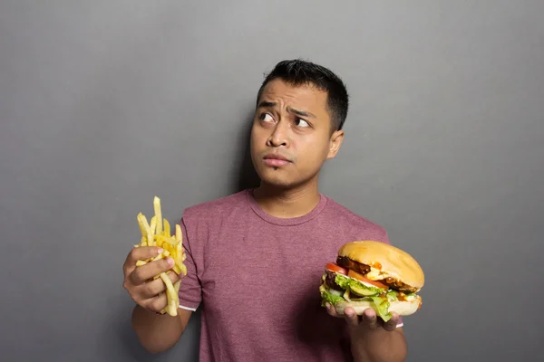 Jeune homme réfléchissant tout en tenant hamburger et frites — Photo