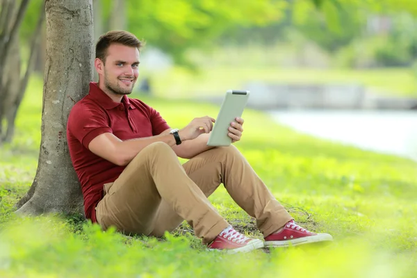 Bir tablet oynarken ağacın altında oturan üniversite öğrencisi — Stok fotoğraf