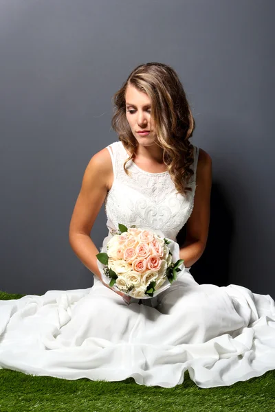 Beautiful young bride sitting on the grass while holding a bouqu — Stock Photo, Image