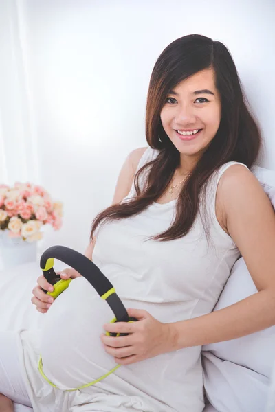 Güzel hamile Asyalı kadın onun bebek şiş kulaklık kullanarak — Stok fotoğraf