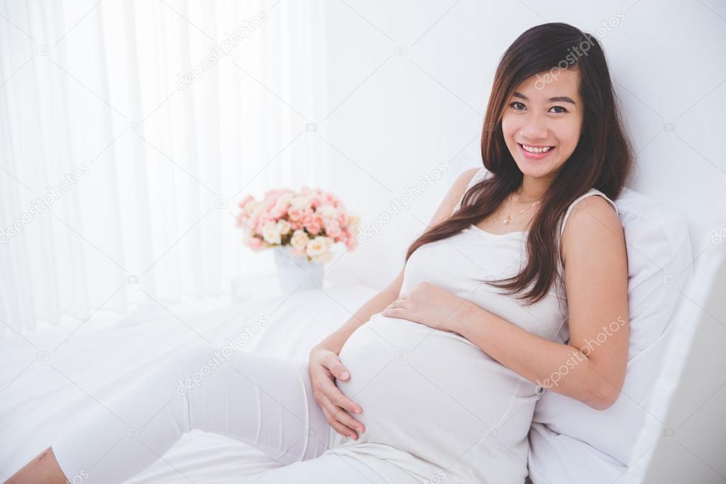 Beautiful pregnant asian woman leaning on a bed, smiling