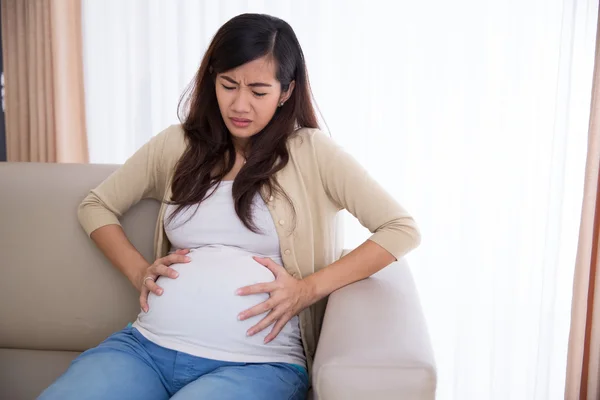 У беременной азиатки болит живот, когда она сидит на диване — стоковое фото